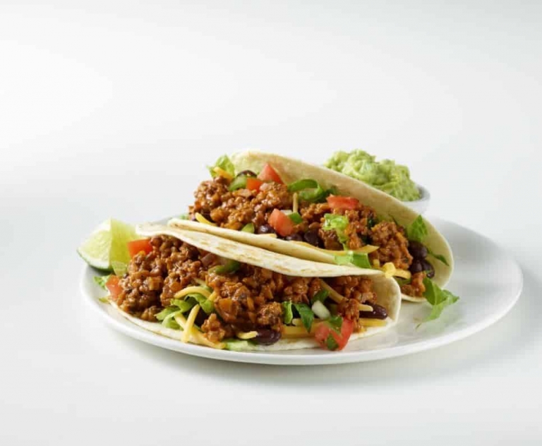 Taco Flavored Walnut Crumbles in flour tortillas plated with black beans, cheese, lettuce, guacamole, and lime wedges.