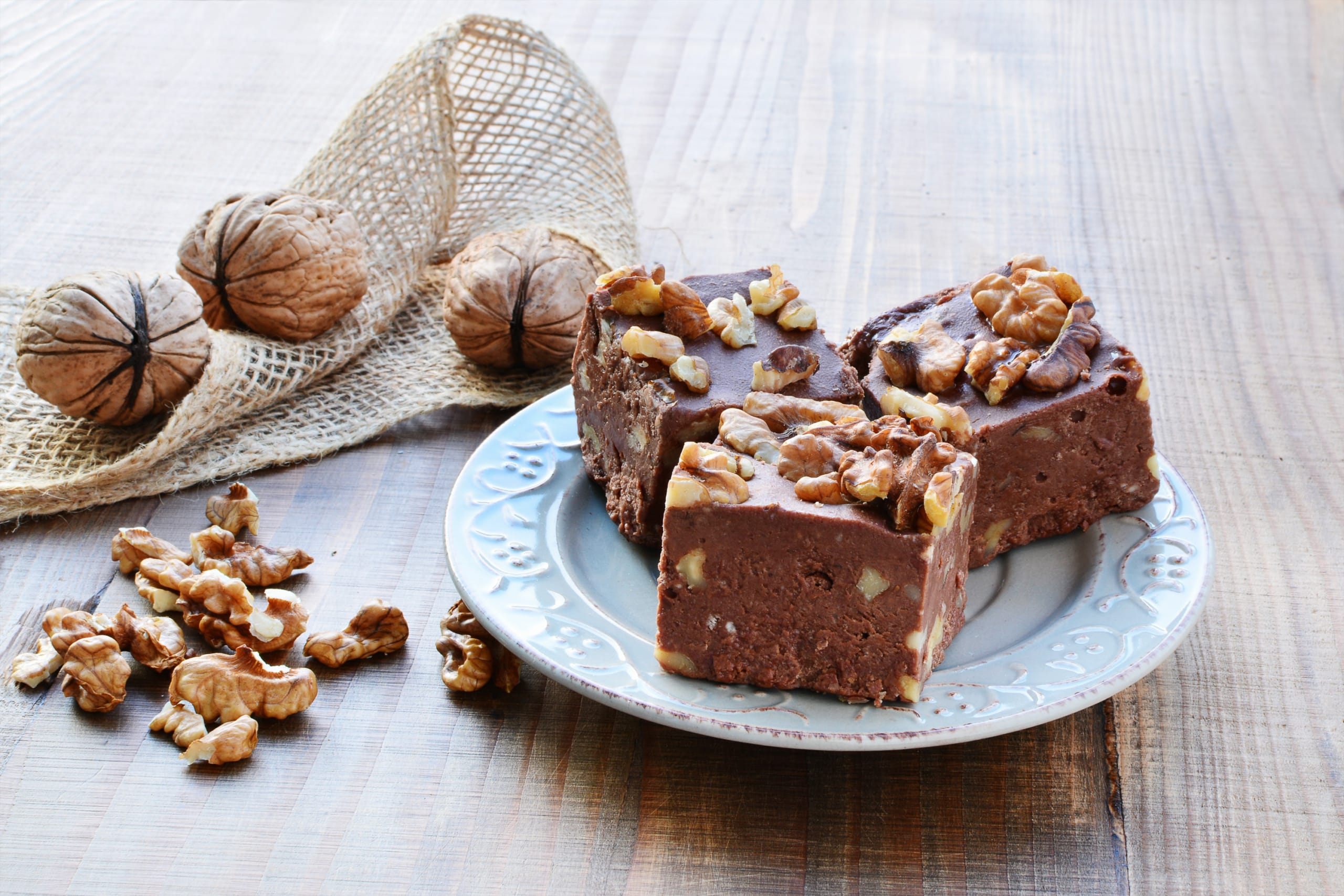 Three pieces of walnut fudge on a plate with bits of walnuts to the left and two whole nuts in the shell.