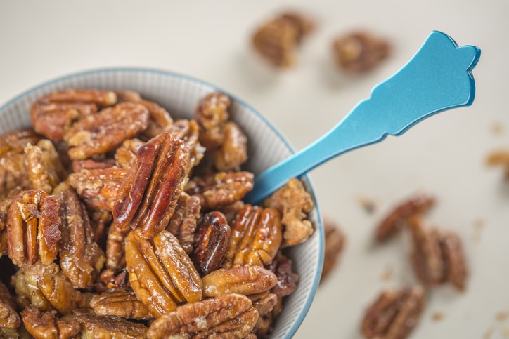 Candied Pecans in a white bowl with a blue spoon and more pecans spilled on the counter.